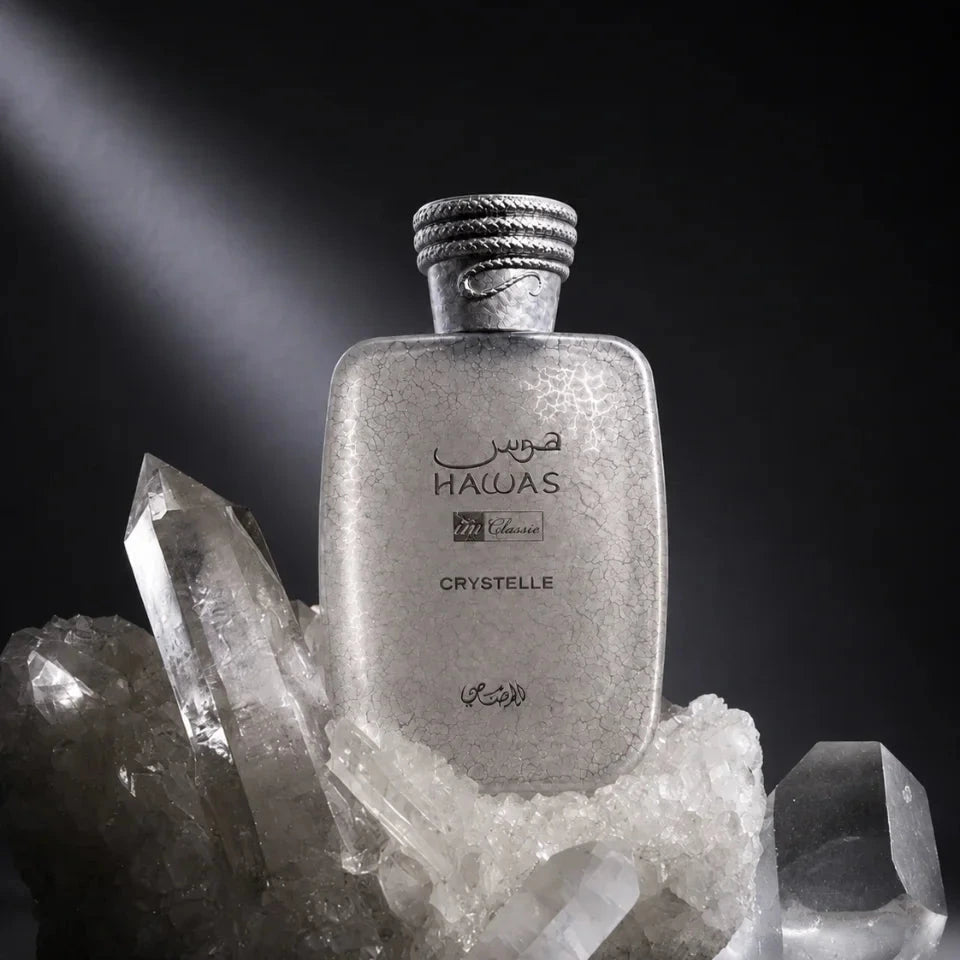 A frosted glass perfume bottle labeled 'HALAS' and 'CRYSTELLE' with Arabic text, styled on a cluster of clear quartz crystals against a dark studio background with dramatic lighting.